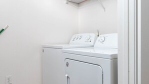 Un appareil compact, blanc, combinant une machine à laver et un sèche-linge à chargement frontal, doté d’un panneau de commande, d’une prise de courant et d’une étagère métallique fixée au mur.