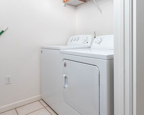 A compact, white front-loading washer and dryer unit with a control panel, a power outlet, and a metal rack on the wall. - Phoenix X 913 3 Bedroom Condo (Orange Beach)