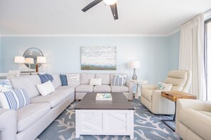 A living room with a sectional sofa, a coffee table, a side table, and a chair. There is a painting on the wall and a ceiling fan. - Phoenix X 513 3 Bedroom Condo (Orange Beach)