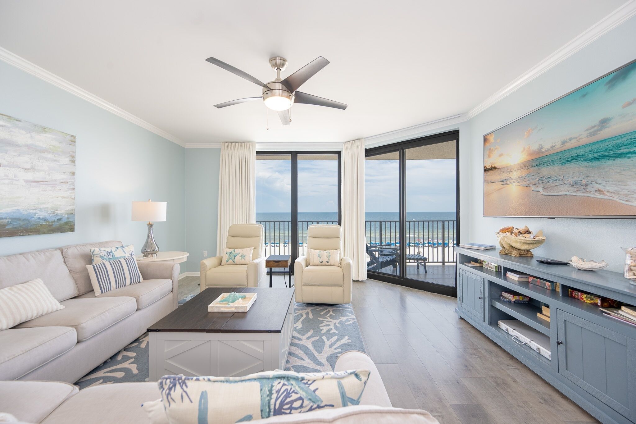 A spacious living room with a sofa, armchairs, a coffee table, and a TV stand. A ceiling fan is visible, and there is a large window offering a view of the beach.