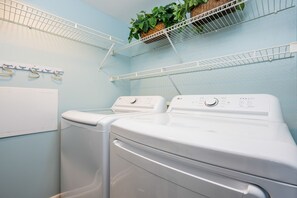 A laundry area with two white front-loading washing machines, a white drying rack, and a wall-mounted drying rack with plants. - Phoenix X 513 3 Bedroom Condo (Orange Beach)