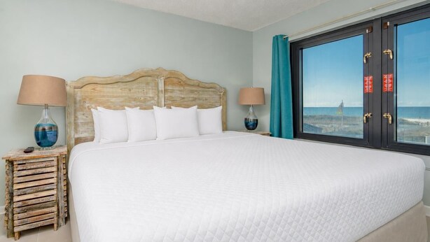 A hotel room with a large bed, two bedside tables, a view of the beach, and a wooden headboard. - Island Winds East 107 2 Bedroom Condo (Gulf Shores)