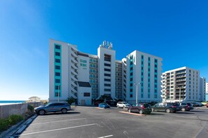 A multi-story hotel with a prominent 'Island Winds East' sign, a parking lot with several cars, and a clear blue sky. - Island Winds East 107 2 Bedroom Condo (Gulf Shores)