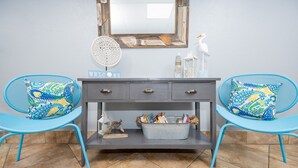 A room with a grey console table, two blue chairs with patterned cushions, a 'WELCOME' sign, a decorative mirror, and a small bird figurine. - Island Winds East 107 2 Bedroom Condo (Gulf Shores)