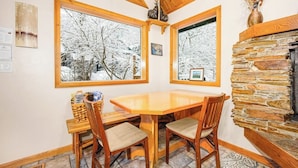 Una habitación acogedora con una mesa y sillas de madera, ventanas amplias con vistas nevadas, una chimenea de piedra y un techo de madera.