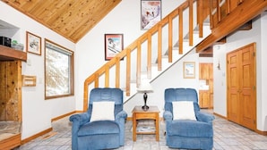Una habitación con una escalera de madera, dos sillones azules, una pequeña mesa con una lámpara y una alfombra estampada.