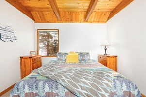 Un dormitorio con techo de madera, una cama grande con ropa de cama estampada, una ventana con vista a árboles y una mesita de noche con lámpara.