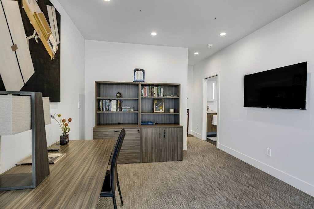 A modern living room with a dining area, a bookshelf, a flat-screen TV mounted on the wall, and a large mirror.