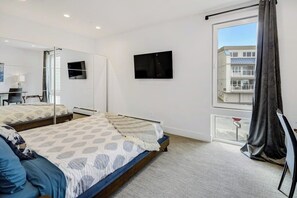 A modern hotel room with a large bed, a flat-screen TV, a window with curtains, and a view of a building outside.