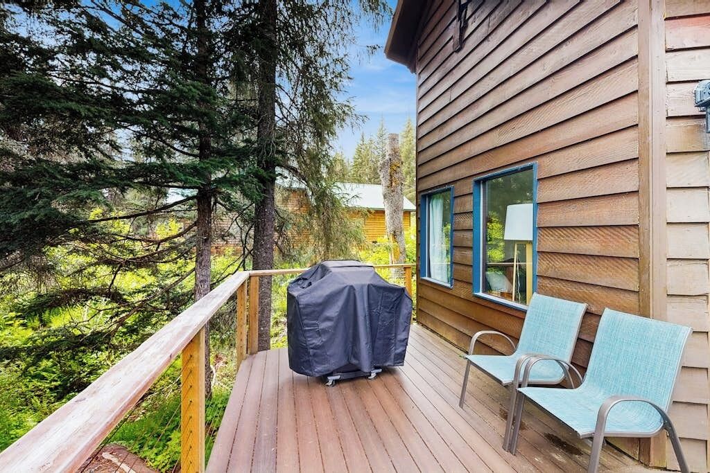A wooden deck with a barbecue grill and two chairs, overlooking a forested area.