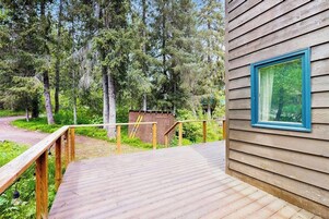 Una terraza de madera que lleva a una cabaña con un ventanal de marco azul, rodeada de un bosque.