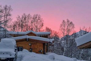 A wooden cabin with a snow-covered roof, surrounded by trees and a pink sky. - Remote 2BR Log Cabin Tucked Outside Anchorage (Eagle River)