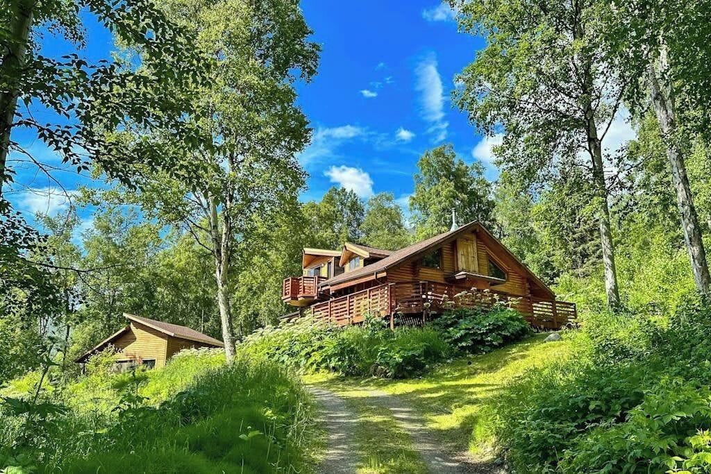 A cabin with a deck and a separate building, surrounded by trees and a clear sky.