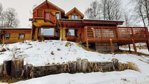 A wooden cabin with multiple levels and large windows, situated on a snowy hill with a wooden fence in the foreground. - Remote 2BR Log Cabin Tucked Outside Anchorage (Eagle River)