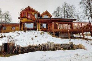 A wooden cabin with multiple levels and large windows, situated on a snowy hill with a wooden fence in the foreground. - Remote 2BR Log Cabin Tucked Outside Anchorage (Eagle River)