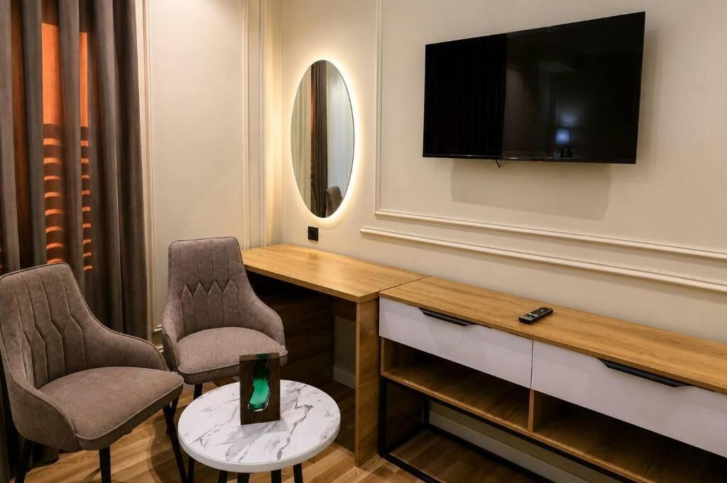 A hotel room with a TV mounted on the wall, a round table with two chairs, a wooden desk with drawers, and a mirror with a light.