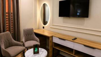 A hotel room with a TV mounted on the wall, a round table with two chairs, a wooden desk with drawers, and a mirror with a light.