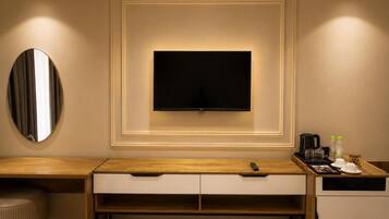 A hotel room with a flat-screen TV mounted on the wall, a wooden desk with drawers, a round mirror, and a tray with a coffee maker and glasses.