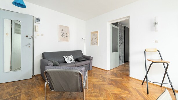 Een kamer met een grijze bank, een kleine houten stoel, een blauwe hanglamp en een deur met een spiegel.