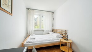Een kleine, net kamer met een enkel bed, een nachtkastje met lamp, een raam met gordijnen en een ingelijst kunstwerk aan de muur.