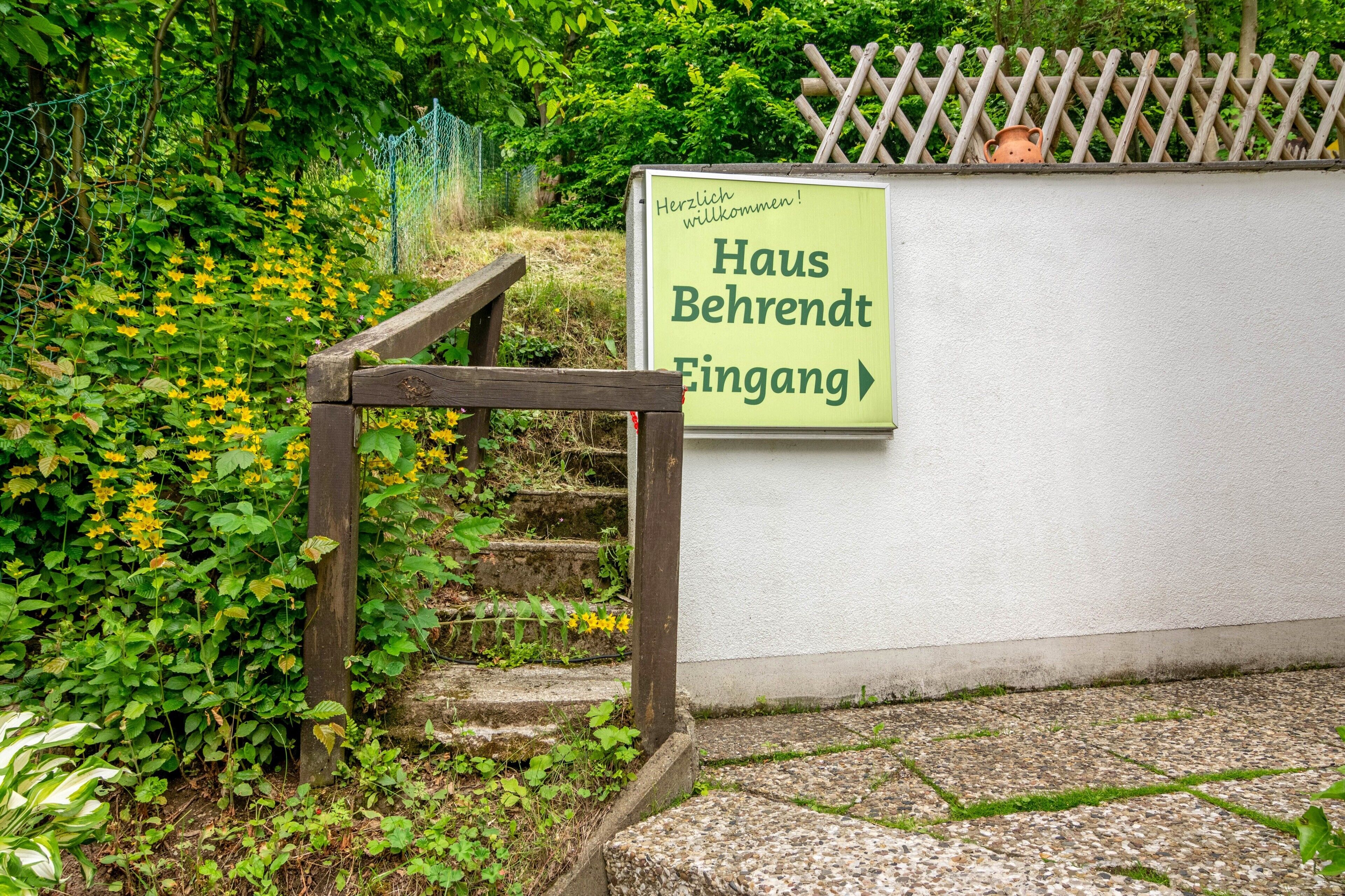 Un escalier en bois mène à l’entrée d’un bâtiment, où se trouve une enseigne indiquant « Haus Behrendt Eingang » ainsi qu’un message de bienvenue.