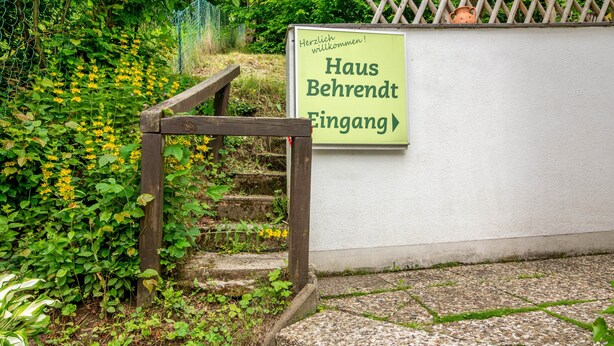 Una escalera de madera que conduce a la entrada de un edificio con un letrero que dice 'Haus Behrendt Eingang' y un mensaje de bienvenida.