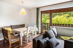 Ein gemütliches Zimmer mit Esstisch, Stühlen, einer Couch und einem großen Fenster mit Blick ins Grüne.
