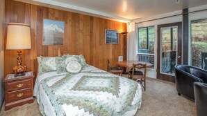 A bedroom with a wooden paneled wall, a bed with a quilt, a nightstand, a desk, and a chair.