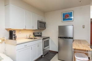 A compact kitchen with white cabinets, a microwave, a stainless steel refrigerator, and a coffee maker. - Slopeside Studio Above The Bake Shop (Girdwood)