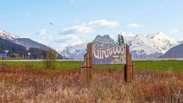 Ein hölzernes Schild mit der Aufschrift 'Welcome to Lindwood Alaska', vor einer Berglandschaft und blauem Himmel.