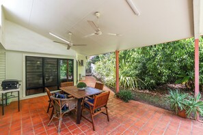 Eine überdachte Terrasse mit Holztisch und -stühlen, einem Deckenventilator und Blick auf einen Garten mit unterschiedlichen Pflanzen.