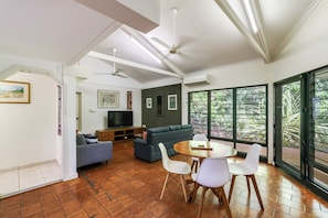 A modern living room with a flat-screen TV, a grey sofa, a dining table with chairs, and large windows with a view of greenery.