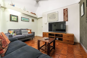 A living room with a grey sofa, a coffee table, a flat-screen TV on a wooden stand, and a wall with framed pictures. - Palms Bungalow Nightcliff with Pool | 2BD, 2BTH (Nightcliff)