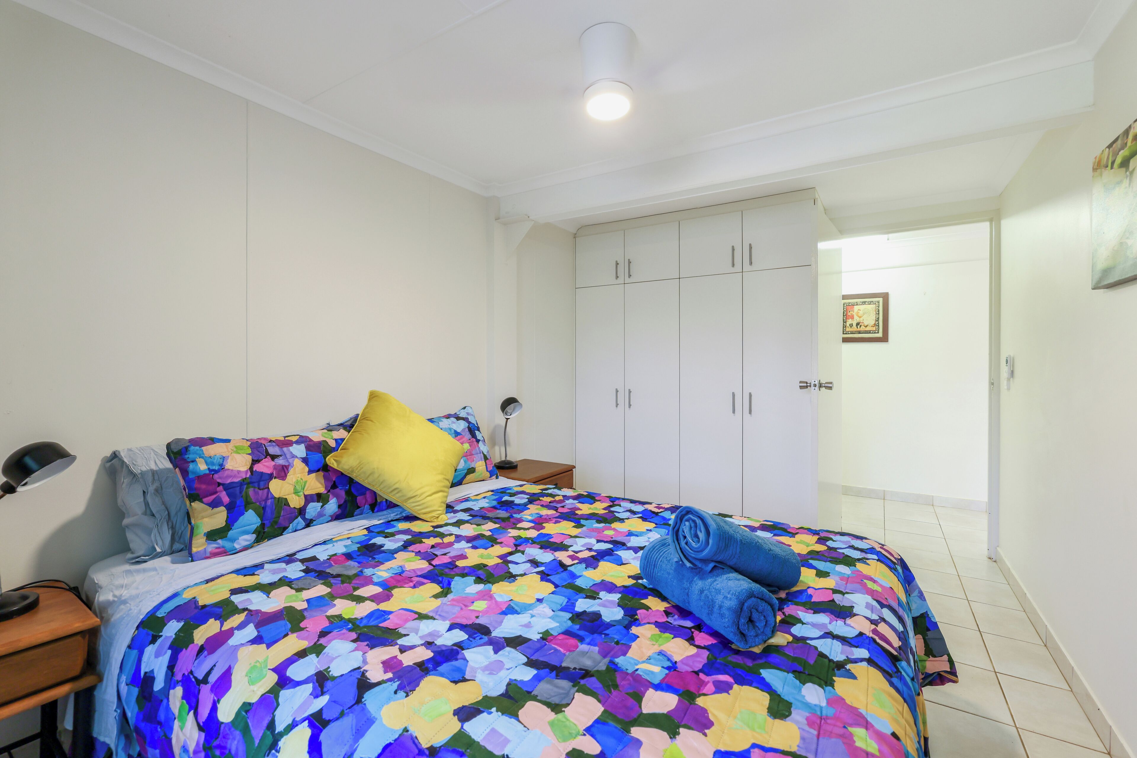 A bedroom with a bed, a nightstand, a lamp, a wardrobe, and a door leading to another room.