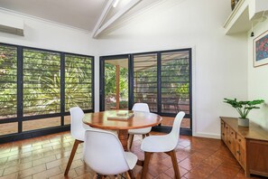 A dining area with a round table, four chairs, and large windows with a view of greenery. - Palms Bungalow Nightcliff with Pool | 2BD, 2BTH (Nightcliff)