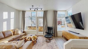 Ein modernes Wohnzimmer mit einer Ledersofa, einem schwarzen Sessel, einem Flachbildfernseher und einem großen Fenster mit Blick auf eine verschneite Landschaft.