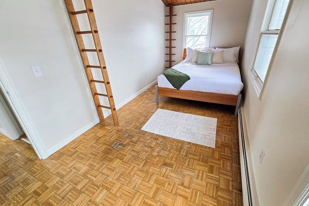 A bedroom with a wooden floor, a bed with pillows, a ladder, and a window.