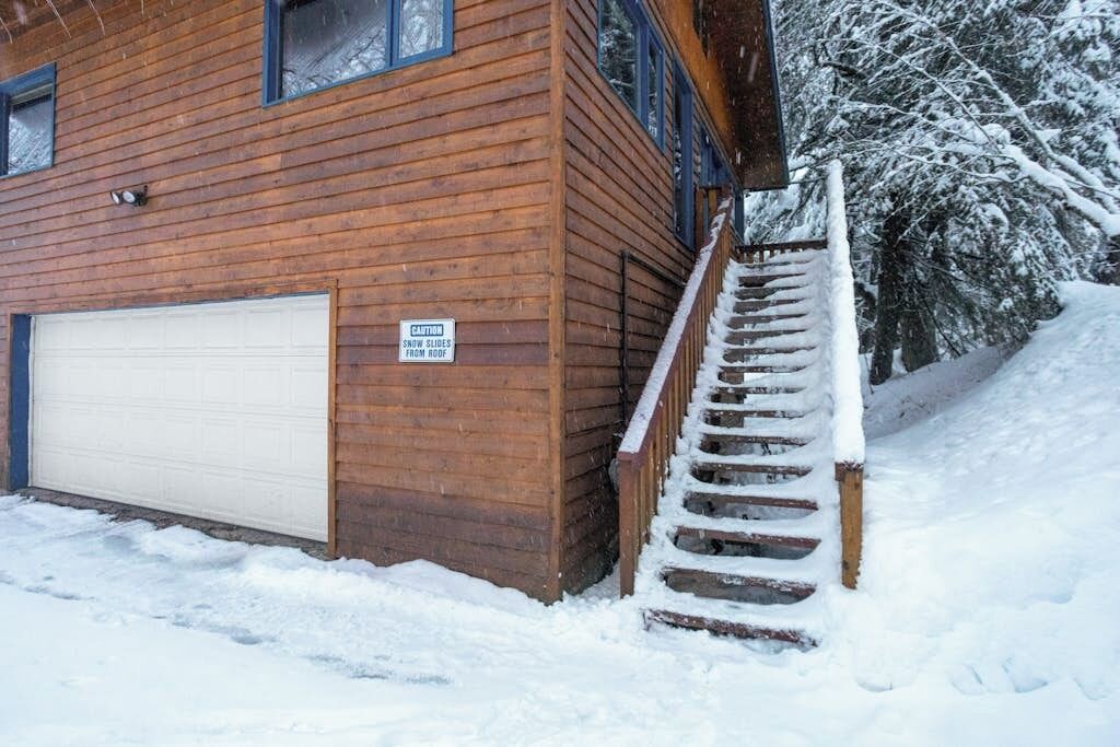 A wooden building with a garage door, a staircase with snow on the steps, and a caution sign about snow slides from the roof.