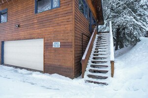 Edificio de madera con puerta de garaje, una escalera con nieve en los peldaños y un letrero de precaución sobre deslizamientos de nieve desde el techo.