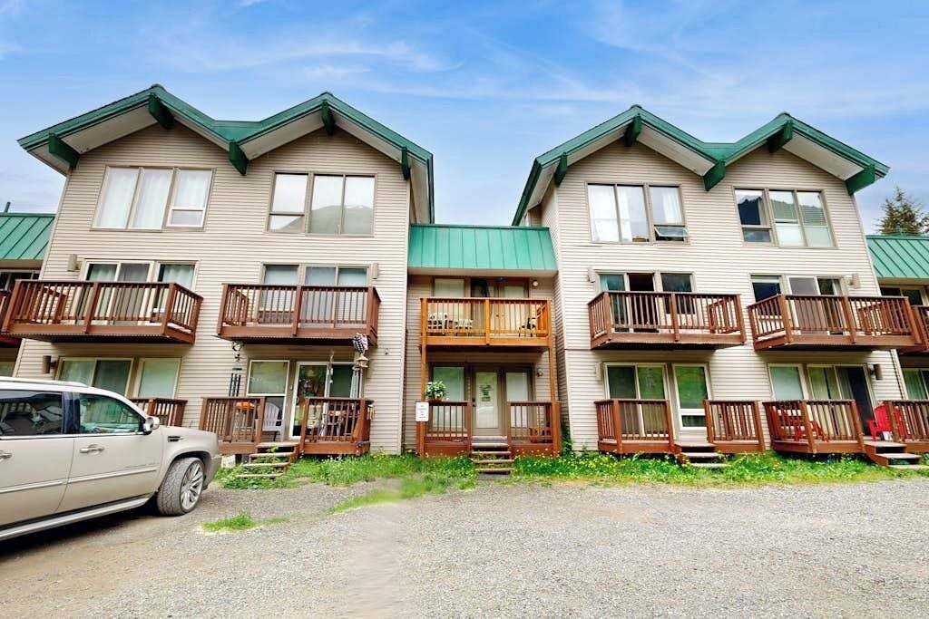 A row of two-story buildings with balconies, a car parked in front, and a gravel driveway.