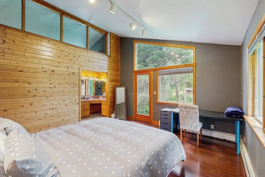 A bedroom with a large bed, a vanity with a mirror, a desk, and a chair. There is a large window providing a view of trees.