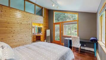 A bedroom with a large bed, a vanity with a mirror, a desk, and a chair. There is a large window providing a view of trees.