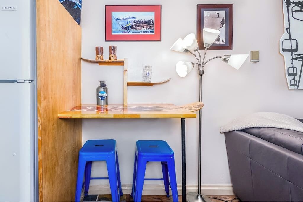 Een compact woongedeelte met een kitchenette, een eethoek met krukken, een bank en wandplanken met decoratieve objecten.