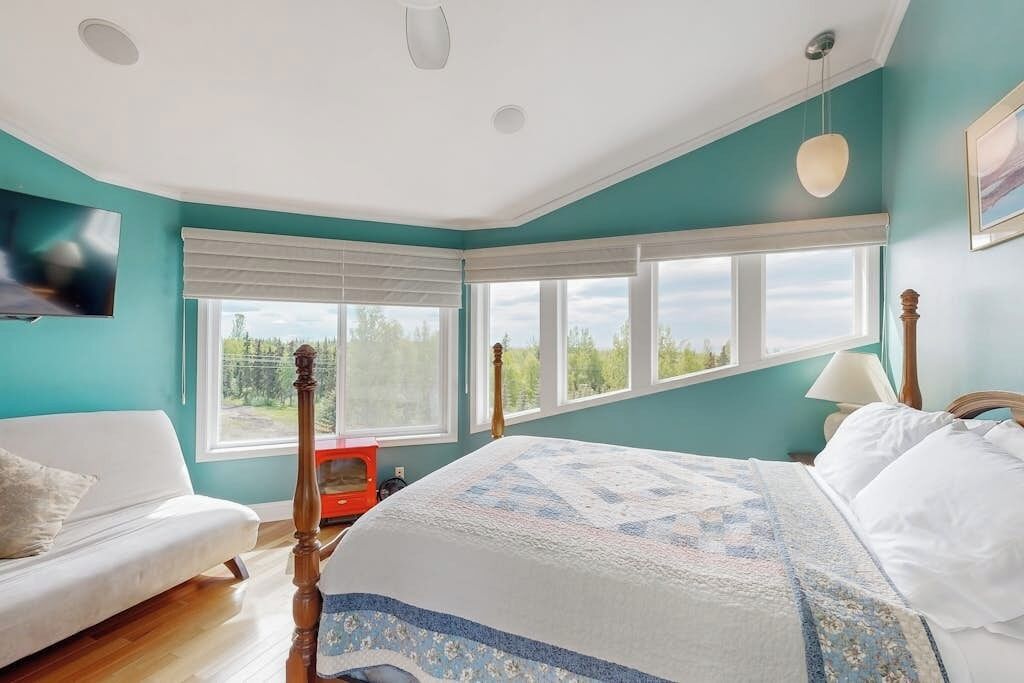 A bedroom with a large bed, a window with blinds, a red chair, and a white sofa.