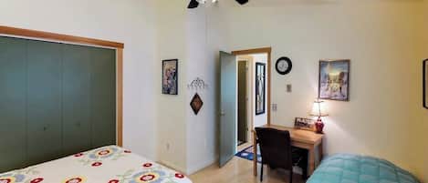 A bedroom with a bed, a desk, a chair, a ceiling fan, and a door leading to another room.
