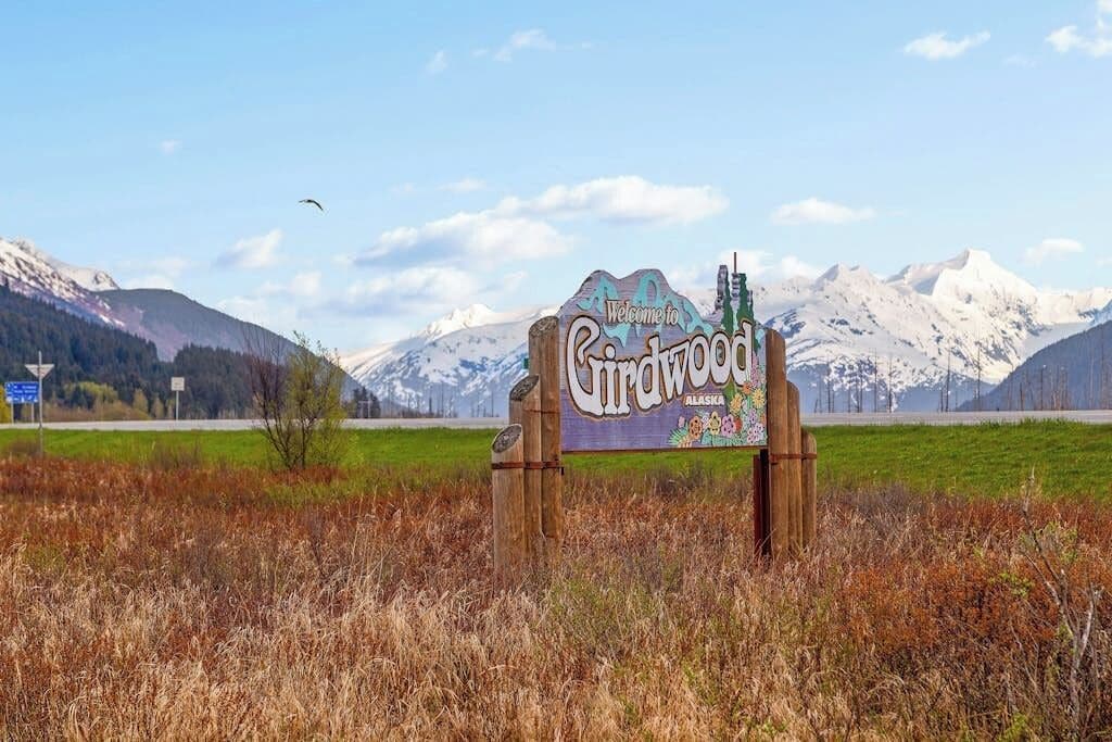 A wooden sign with 'Welcome to Lindwood Alaska' written on it, set against a backdrop of mountains and a clear sky.