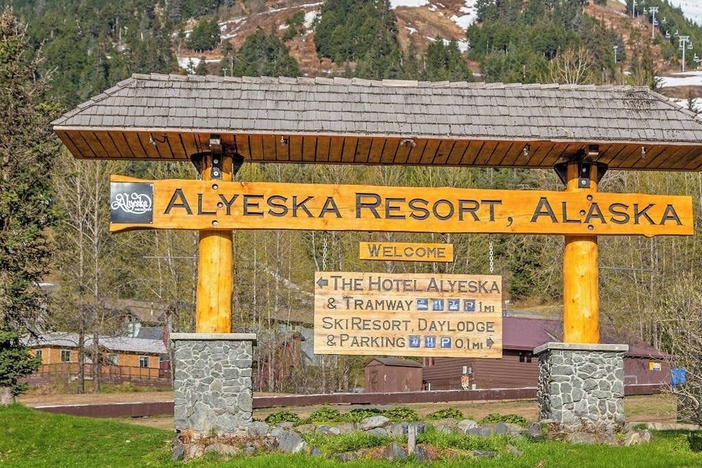 A wooden sign with 'Alyeska Resort, Alaska' and directional arrows pointing to 'The Hotel Alyeska & Tramway' and 'Skiresort, Daylodge & Parking.'