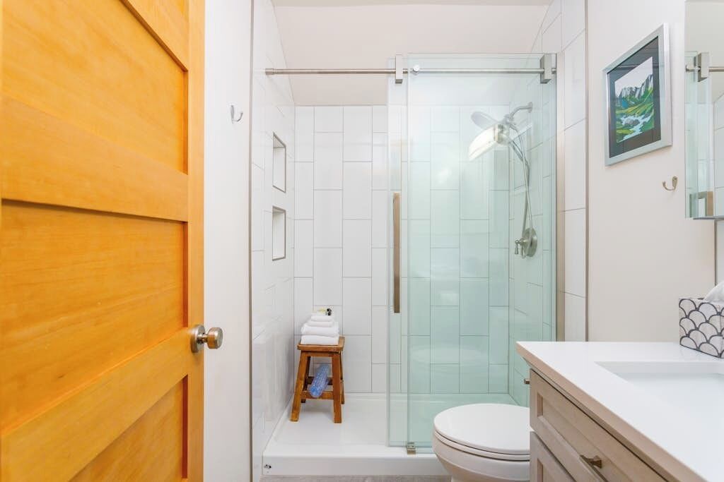 A bathroom with a glass shower enclosure, a wooden door, a white toilet, and a small wooden stool with towels.