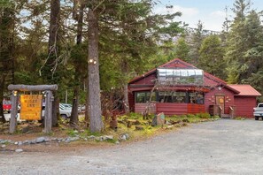 A rustic cabin with a red exterior and a glass front, surrounded by trees and a gravel path.