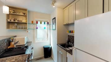A compact kitchen with a gas stove, a sink, and a refrigerator. There is a window with a view of the outside, and a trash bin is visible.